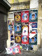 A wall covered with a collage of various posters featuring different designs and advertisements. The posters predominantly advertise 'Craft Cocktails' with artistic portraits and vibrant text, suggesting themes of nightlife or beverages. Some of the posters are torn or overlapped, showing signs of wear and urban decay. The wall itself is weathered, with visible graffiti above the posters.