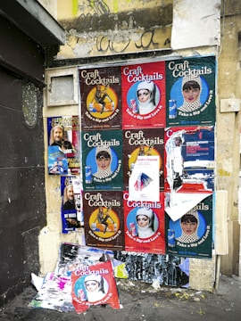 A wall covered with a collage of various posters featuring different designs and advertisements. The posters predominantly advertise 'Craft Cocktails' with artistic portraits and vibrant text, suggesting themes of nightlife or beverages. Some of the posters are torn or overlapped, showing signs of wear and urban decay. The wall itself is weathered, with visible graffiti above the posters.