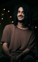 A person with long, curly hair is sitting in a relaxed pose with a gentle smile and closed eyes. The lighting is soft, casting warm tones. In the background, several small, blurred lights create a cozy, intimate atmosphere.