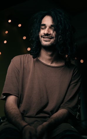 A person with long, curly hair is sitting in a relaxed pose with a gentle smile and closed eyes. The lighting is soft, casting warm tones. In the background, several small, blurred lights create a cozy, intimate atmosphere.