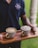 A person holds a wooden tray containing two intricately designed coffee cups. Each cup features a distinct pattern and has a decorative handle. The surfaces of the drinks display elegant latte art. The person wears a navy blue shirt with an emblem, standing against a blurred green backdrop.