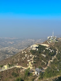 A mountainous landscape with a telecommunications tower atop a hill. The hill is covered with rocks, sparse vegetation, and a few trees. A small pavilion is located at the base of the hill, and a pathway leads up the slope. The sky is clear and blue.