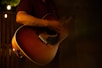 A person is playing an acoustic guitar in a dimly lit setting, with warm lighting creating a cozy and intimate atmosphere. The focus is on the guitar and the player's hands, suggesting a musical performance or practice session.