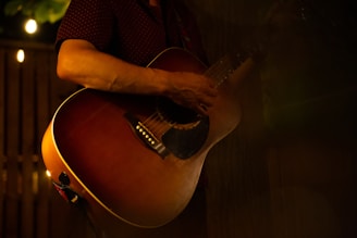 A person is playing an acoustic guitar in a dimly lit setting, with warm lighting creating a cozy and intimate atmosphere. The focus is on the guitar and the player's hands, suggesting a musical performance or practice session.