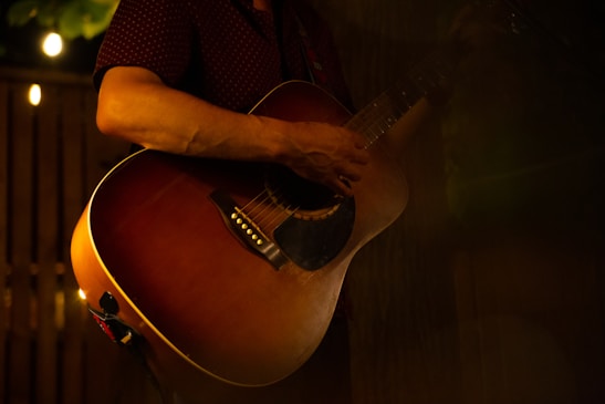 A person is playing an acoustic guitar in a dimly lit setting, with warm lighting creating a cozy and intimate atmosphere. The focus is on the guitar and the player's hands, suggesting a musical performance or practice session.