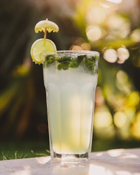 A refreshing glass of lemonade garnished with mint leaves, a slice of lime, and a small decorative umbrella. The background is softly blurred with green and yellow hues, suggesting an outdoor setting with natural light.