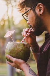 A person with glasses sips from a fresh coconut using a striped straw, while holding it with both hands. The coconut has a small decorative umbrella. The scene is set outdoors with a blurred background of greenery and warm, soft lighting.