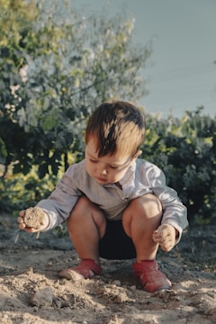 A child crouches on the ground, intently examining a clump of dirt in their hand. The child is wearing a striped shirt and red shoes, and is surrounded by a natural setting with greenery and a clear sky in the background.