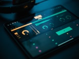 A tablet screen displays a music production software interface with various controls like dials, sliders, and waveform visuals. The background is dark, with illuminated elements in yellow and teal hues. Nearby, a pair of headphones lies on a flat surface.