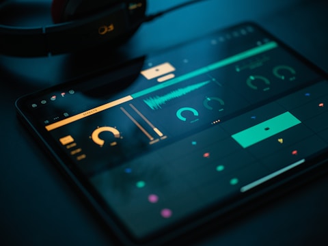 A tablet screen displays a music production software interface with various controls like dials, sliders, and waveform visuals. The background is dark, with illuminated elements in yellow and teal hues. Nearby, a pair of headphones lies on a flat surface.