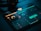 A tablet screen displays a music production software interface with various controls like dials, sliders, and waveform visuals. The background is dark, with illuminated elements in yellow and teal hues. Nearby, a pair of headphones lies on a flat surface.