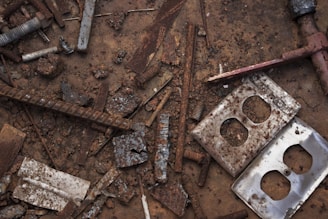 A collection of rusted metal objects and debris scattered across a dirt-covered surface. The items include corroded screws, nuts, a bent rebar, and several pieces of broken metal plates, including a couple of switch plates.