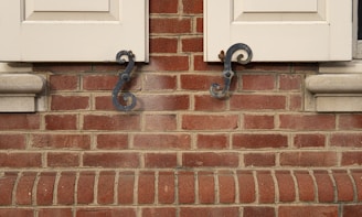 A close-up view of a red brick wall featuring two wooden window shutters partially open. The shutters are accented with ornate, black metal hardware in a swirling design. The bricks are aligned in a uniform pattern with visible mortar between them.