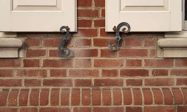 A close-up view of a red brick wall featuring two wooden window shutters partially open. The shutters are accented with ornate, black metal hardware in a swirling design. The bricks are aligned in a uniform pattern with visible mortar between them.