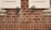 A close-up view of a red brick wall featuring two wooden window shutters partially open. The shutters are accented with ornate, black metal hardware in a swirling design. The bricks are aligned in a uniform pattern with visible mortar between them.