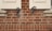 A close-up view of a red brick wall featuring two wooden window shutters partially open. The shutters are accented with ornate, black metal hardware in a swirling design. The bricks are aligned in a uniform pattern with visible mortar between them.