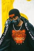 A person is wearing a black beanie hat with a Givenchy Paris logo, a black face mask, a black t-shirt with an orange tiger print and the words Kenzo Paris, a silver chain necklace, and a black jacket with white text along the sleeves.