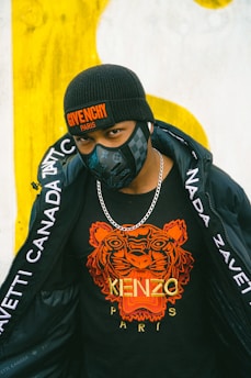 A person is wearing a black beanie hat with a Givenchy Paris logo, a black face mask, a black t-shirt with an orange tiger print and the words Kenzo Paris, a silver chain necklace, and a black jacket with white text along the sleeves.