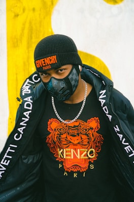A person is wearing a black beanie hat with a Givenchy Paris logo, a black face mask, a black t-shirt with an orange tiger print and the words Kenzo Paris, a silver chain necklace, and a black jacket with white text along the sleeves.