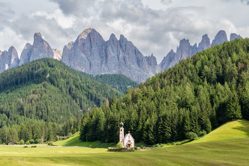 a small white building in a grassy field with trees and mountains in the background with Dolomites in the background