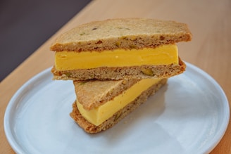 A delicious-looking ice cream sandwich composed of vibrant yellow mango ice cream between two thick, homemade cookies with visible pieces of pistachio. The dessert is neatly placed on a simple white plate, enhancing its appetizing presentation. The setting includes a wooden surface, adding a rustic touch.