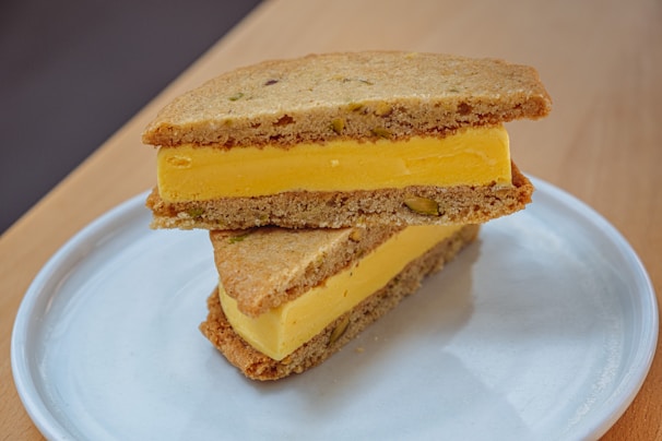 A delicious-looking ice cream sandwich composed of vibrant yellow mango ice cream between two thick, homemade cookies with visible pieces of pistachio. The dessert is neatly placed on a simple white plate, enhancing its appetizing presentation. The setting includes a wooden surface, adding a rustic touch.