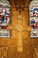 A richly decorated church interior featuring ornate golden patterns and intricate stained glass windows depicting religious figures. A prominent cross with detailed engravings is centrally placed against a backing of elaborate designs, showcasing Christian iconography.