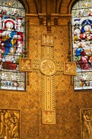 A richly decorated church interior featuring ornate golden patterns and intricate stained glass windows depicting religious figures. A prominent cross with detailed engravings is centrally placed against a backing of elaborate designs, showcasing Christian iconography.