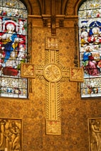 A richly decorated church interior featuring ornate golden patterns and intricate stained glass windows depicting religious figures. A prominent cross with detailed engravings is centrally placed against a backing of elaborate designs, showcasing Christian iconography.