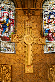 A richly decorated church interior featuring ornate golden patterns and intricate stained glass windows depicting religious figures. A prominent cross with detailed engravings is centrally placed against a backing of elaborate designs, showcasing Christian iconography.