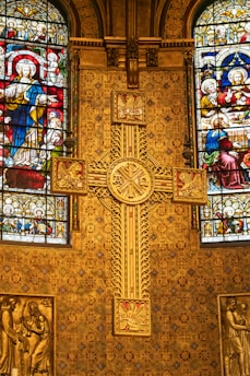 A richly decorated church interior featuring ornate golden patterns and intricate stained glass windows depicting religious figures. A prominent cross with detailed engravings is centrally placed against a backing of elaborate designs, showcasing Christian iconography.