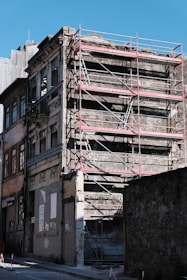 A dilapidated building is undergoing restoration, with scaffolding surrounding its facade. The structure appears aged and worn, with several floors visible. Adjacent buildings also exhibit old architecture, and a clear blue sky is in the background.