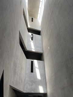 A tall, narrow interior space with concrete walls featuring minimalistic design and sharp geometric lines. Natural light streams in from a skylight above, casting distinct rectangular shadows and highlights on the walls. The structure includes protruding rectangular elements with small vertical windows.