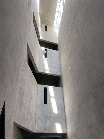 A tall, narrow interior space with concrete walls featuring minimalistic design and sharp geometric lines. Natural light streams in from a skylight above, casting distinct rectangular shadows and highlights on the walls. The structure includes protruding rectangular elements with small vertical windows.