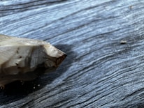 A pointed, polished crystal or mineral specimen lies on a textured dark wooden surface. The sharp tip and multi-faceted nature of the mineral are prominent, with subtle reflections and a mix of dark and light tones on the crystal.
