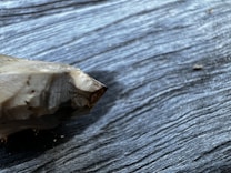 A pointed, polished crystal or mineral specimen lies on a textured dark wooden surface. The sharp tip and multi-faceted nature of the mineral are prominent, with subtle reflections and a mix of dark and light tones on the crystal.