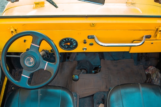 An interior view of a vehicle showing the dashboard and steering wheel. The dashboard is prominently colored in vibrant yellow, with a simple design featuring a speedometer and a metal handle. The steering wheel is black with subtle pink accents, having a classic and straightforward design. The floor has rubber mats and the seats appear to be basic and functional.