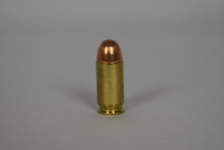 A single bullet standing upright on a plain surface, featuring a metallic casing with a copper-colored tip. The scene is minimalistic with no additional elements or background details.