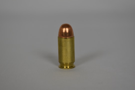 A single bullet standing upright on a plain surface, featuring a metallic casing with a copper-colored tip. The scene is minimalistic with no additional elements or background details.