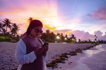 A person with sunglasses and a white shirt stands on a sandy beach, engrossed in a smartphone, against a vibrant sunset sky. Palm trees line the horizon and a few people are scattered in the background near the shoreline. The sky exhibits a gradient of pink, purple, and orange hues, reflecting on the calm water and lending a serene atmosphere.