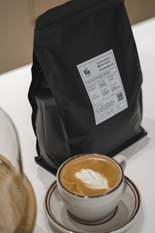 A dark coffee bag with a label sits on a counter next to a cappuccino in a light-colored cup and saucer. The coffee has a latte art design in the foam on top.