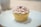 A single cupcake with a swirl of light pink frosting is set on a white plate. The cupcake is wrapped in a shiny, golden paper liner and topped with a small dollop of berry jam.