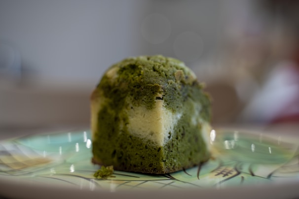 A piece of green and white marble cake sits on a decorative plate with an artistic pattern. The cake has a moist texture and a visually contrasting swirl of colors.