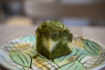 A slice of green matcha cake with a moist texture is placed on a decorative plate. The cake appears to have a marbled pattern with green and cream colors. The plate has a geometric pattern with shades of green and yellow, creating an artistic backdrop for the dessert.