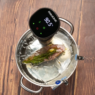 A sous vide device with a digital display showing 90.5°C is attached to a pot of water. Inside the pot, a vacuum-sealed bag contains a piece of meat, herbs, and spices.