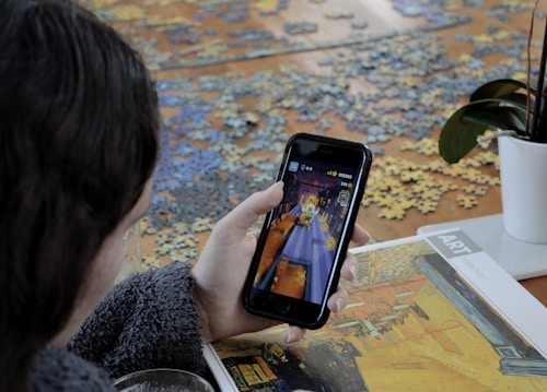 A person is playing a game on a smartphone. In the foreground, there is a puzzle spread out on the table with some pieces assembled, and a booklet featuring artwork. A plant with green leaves is placed on the table.