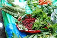 A variety of fresh produce displayed on a table covered with a bright blue and green tarp. A red tray holds an abundance of small red berries surrounded by vibrant green leaves. Long green vegetables are placed next to leafy greens, while carrots and green peppers are arranged in the upper part of the image.