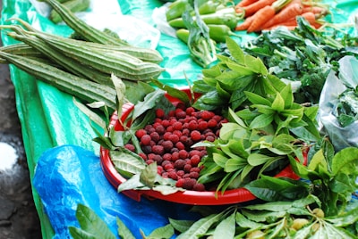 A variety of fresh produce displayed on a table covered with a bright blue and green tarp. A red tray holds an abundance of small red berries surrounded by vibrant green leaves. Long green vegetables are placed next to leafy greens, while carrots and green peppers are arranged in the upper part of the image.