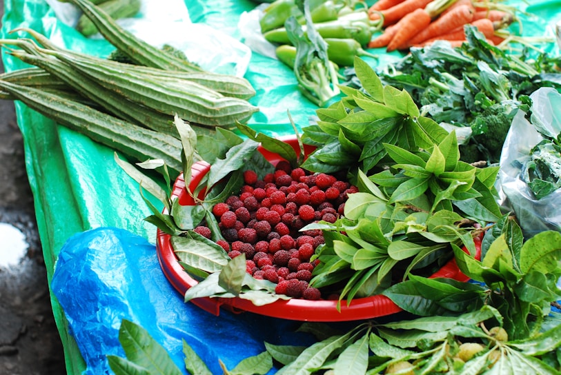 A variety of fresh produce displayed on a table covered with a bright blue and green tarp. A red tray holds an abundance of small red berries surrounded by vibrant green leaves. Long green vegetables are placed next to leafy greens, while carrots and green peppers are arranged in the upper part of the image.