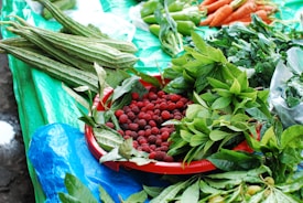 A variety of fresh produce displayed on a table covered with a bright blue and green tarp. A red tray holds an abundance of small red berries surrounded by vibrant green leaves. Long green vegetables are placed next to leafy greens, while carrots and green peppers are arranged in the upper part of the image.